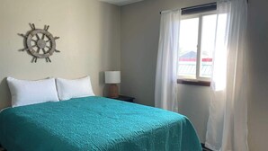 Una habitación con una cama, un timón de barco en la pared, una ventana con cortinas y una mesita de noche con una lámpara.