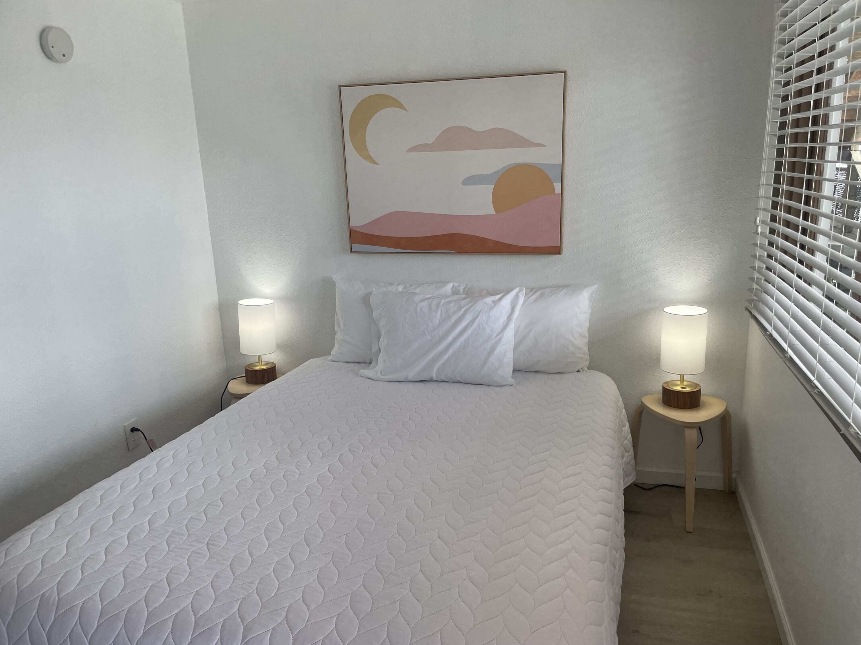 A neatly made bed with a quilted white cover, two bedside lamps, and a wall-mounted painting of a sunset.