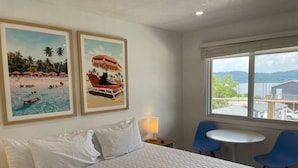 Een hotelkamer met een bed, twee ingelijste foto’s van het strand, een raam met uitzicht, een tafel en twee stoelen.