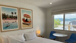 A hotel room with a bed, two framed beach pictures, a window with a view, a table, and two chairs. - Single King Room (Upper Level) (Lake City)