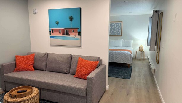 A modern living room with a grey sofa, red patterned pillows, and a painting of a building and palm trees.
