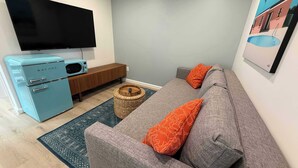 A modern living room with a flat-screen TV, a vintage blue Galanz refrigerator, a wooden cabinet, a grey sofa with orange pillows, and a blue patterned rug.