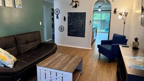 Un salon avec un canapé en cuir marron, une table basse en bois, un fauteuil bleu et une petite table sur laquelle se trouve un ananas. Les murs sont décorés avec un crâne, une roue et un tableau noir orné de dessins.