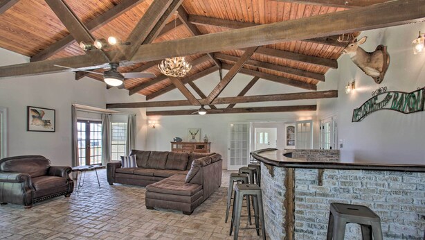 Amplia sala de estar con piso de piedra, un sofá seccional marrón grande, sillones de cuero, techo de madera con vigas a la vista, una araña y una cabeza de venado montada.