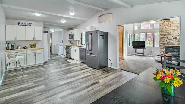 A modern kitchen with white cabinets, a stainless steel refrigerator, and a dining area with a wooden table and a vase of flowers. - 2 Mi to Rainbow Lake: Woodsy Retreat w/ Deck! (Lakeside)
