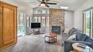 A spacious living room with a stone fireplace, a ceiling fan, and a large window offering a view of the outdoors. - 2 Mi to Rainbow Lake: Woodsy Retreat w/ Deck! (Lakeside)