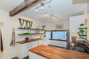 Eine moderne Küche mit Holzarbeitsplatten, weißen Schränken und einer großen Kochinsel. Es gibt Hängeleuchten, ein Fenster mit Jalousien und verschiedene Küchengeräte.