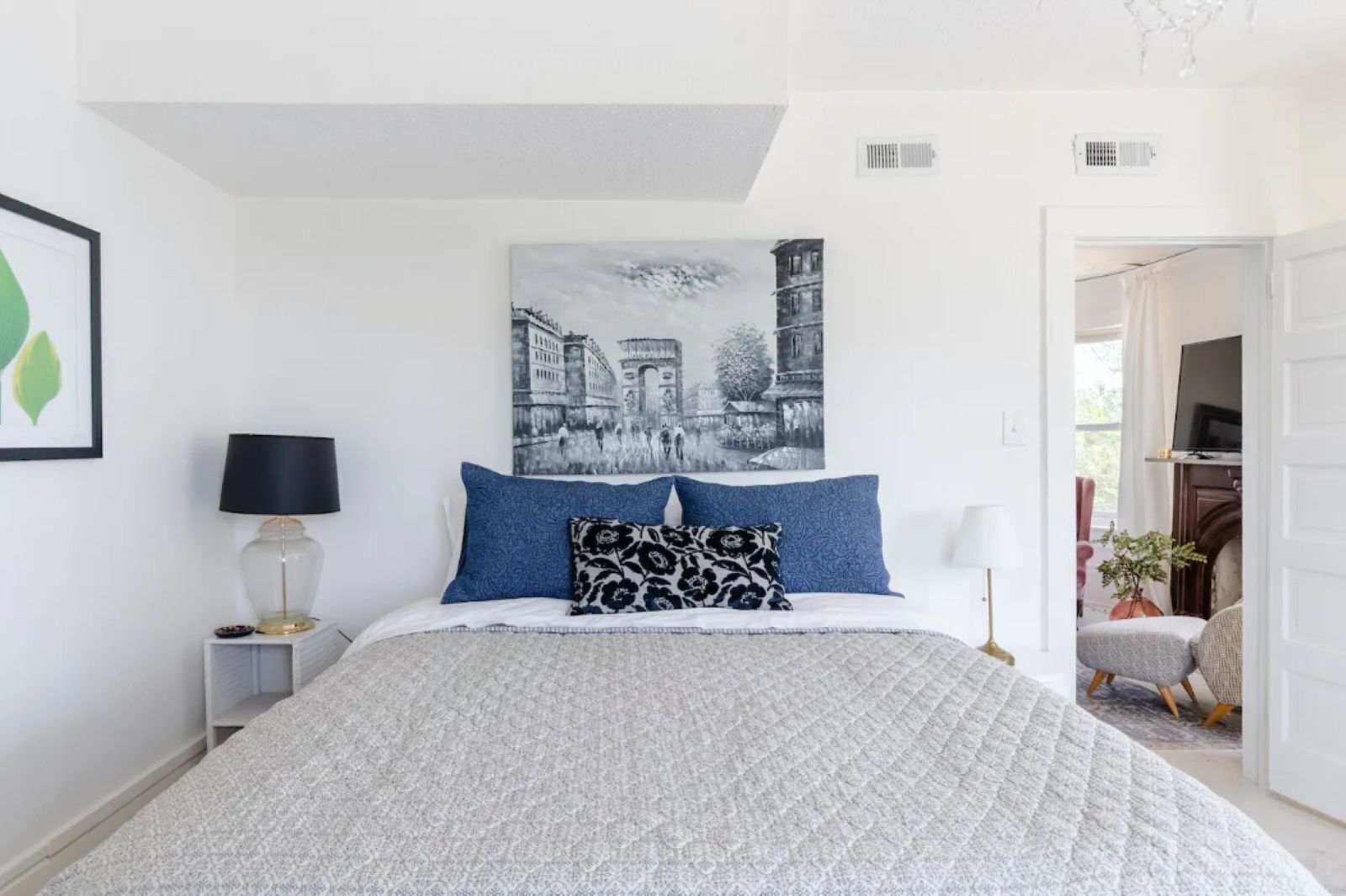 Une chambre à coucher comprenant un lit, une table de chevet, une lampe, un tableau accroché au mur et une porte donnant sur une autre pièce.