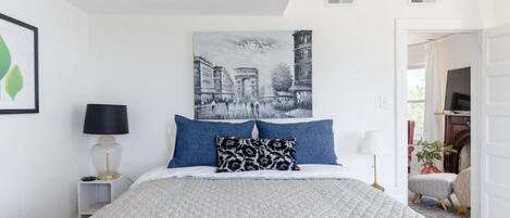 Une chambre à coucher comprenant un lit, une table de chevet, une lampe, un tableau accroché au mur et une porte donnant sur une autre pièce.