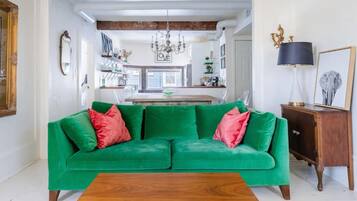 Um sofá verde com almofadas vermelhas, uma mesa de centro de madeira e um candelabro numa sala de estar com cozinha ao fundo.