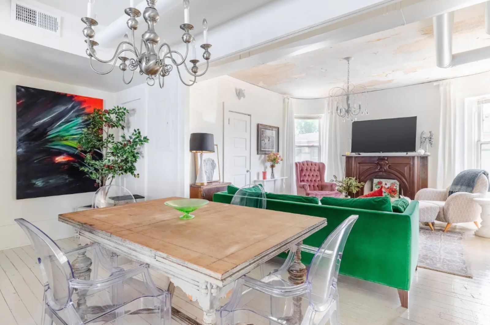 Uma sala de estar ampla com uma mesa de jantar de madeira, cadeiras transparentes, um sofá verde e um lustre.
