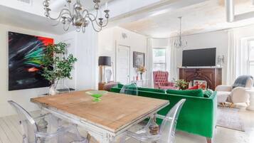 Uma sala de estar ampla com uma mesa de jantar de madeira, cadeiras transparentes, um sofá verde e um lustre.
