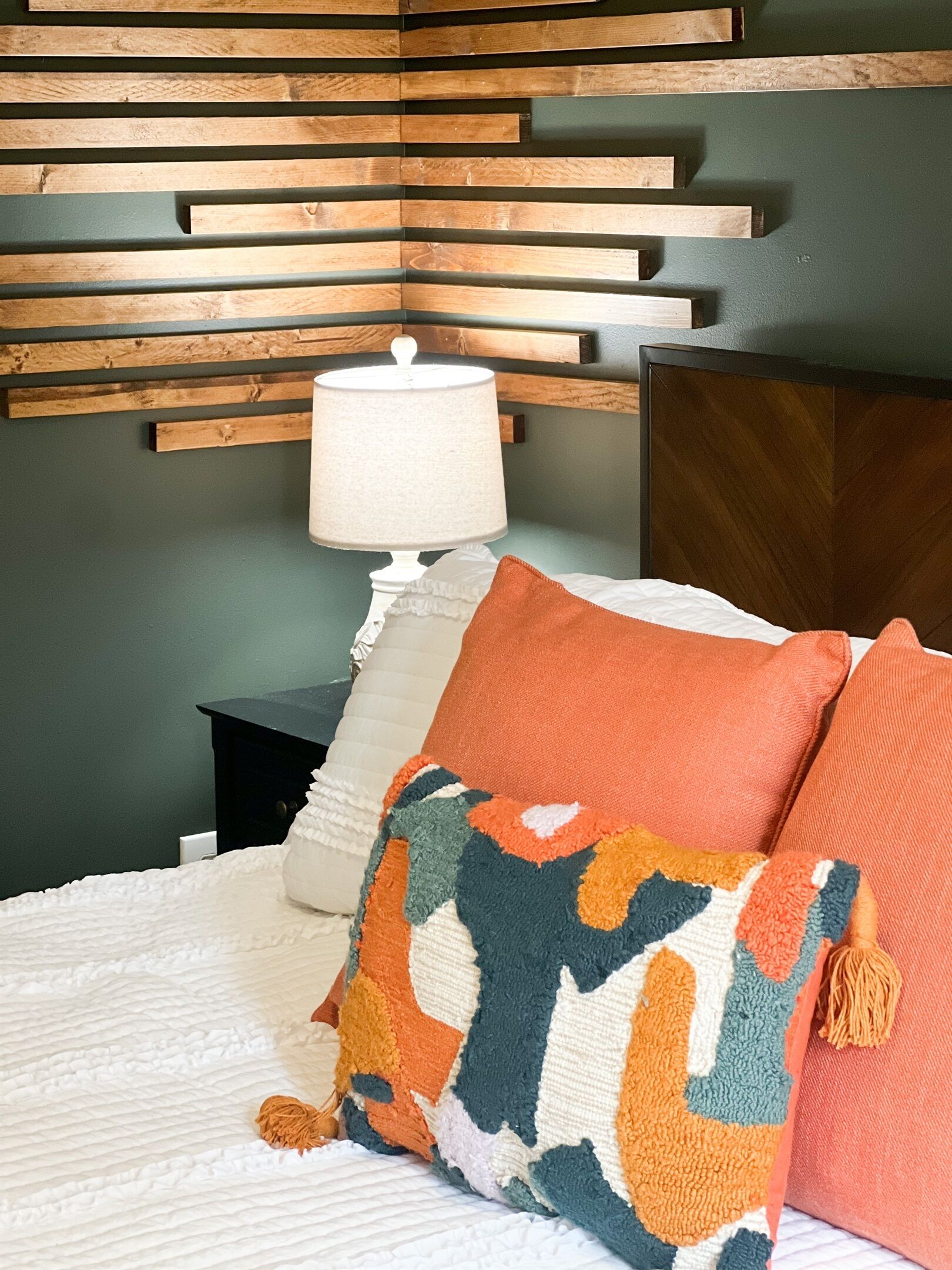 A neatly made bed with a white comforter and two colorful pillows, a bedside table with a white lamp, and a wooden headboard with horizontal slats.
