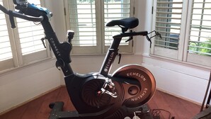 Un vélo d’appartement doté d’une selle et de pédales noires, placé dans une pièce au plancher en bois et aux grandes fenêtres équipées de stores.