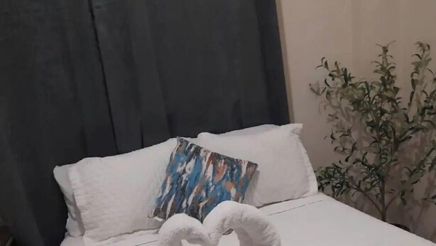 Ein Bett mit weißer gesteppter Bettdecke, einer herzförmigen Handtuchanordnung, einem einzelnen weißen Kopfkissen und einem farbenfrohen Kissen.