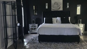 Una camera da letto con un letto grande, una testiera nera, un pavimento in legno, un supporto nero in metallo e un tappeto bianco.