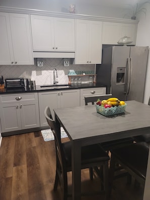 A modern kitchen with white cabinets, a stainless steel refrigerator, a built-in oven, and a dining table with a fruit basket.