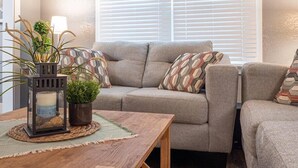 Ein gemütliches Wohnzimmer mit grauem Sofa, gemusterten Kissen, einem Holztisch und einer Topfpflanze.