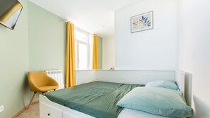 Ein Schlafzimmer mit Bett, Sessel, Heizkörper, einem Fenster mit Vorhängen und einem Wandbild.