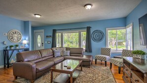Ein Wohnzimmer mit einer braunen Ledersofa, einem Glastisch und gemusterten Teppichen. Zwei Fenster mit Jalousien, eine Uhr und ein Willkommensschild sind ebenfalls vorhanden.