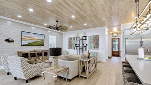 A modern living room with a sofa, a coffee table, and a TV. There is a kitchen area with a refrigerator and a bar counter. - Lucky Catch: Free golf cart, Walk to beach, Heated pool behind house, Game room (Miramar Beach)