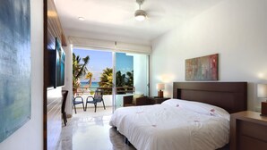 Ein Hotelzimmer mit einem großen Bett, einem Schreibtisch, einem Sessel, einem Fernseher und einem Balkon mit Blick auf Palmen und das Meer.
