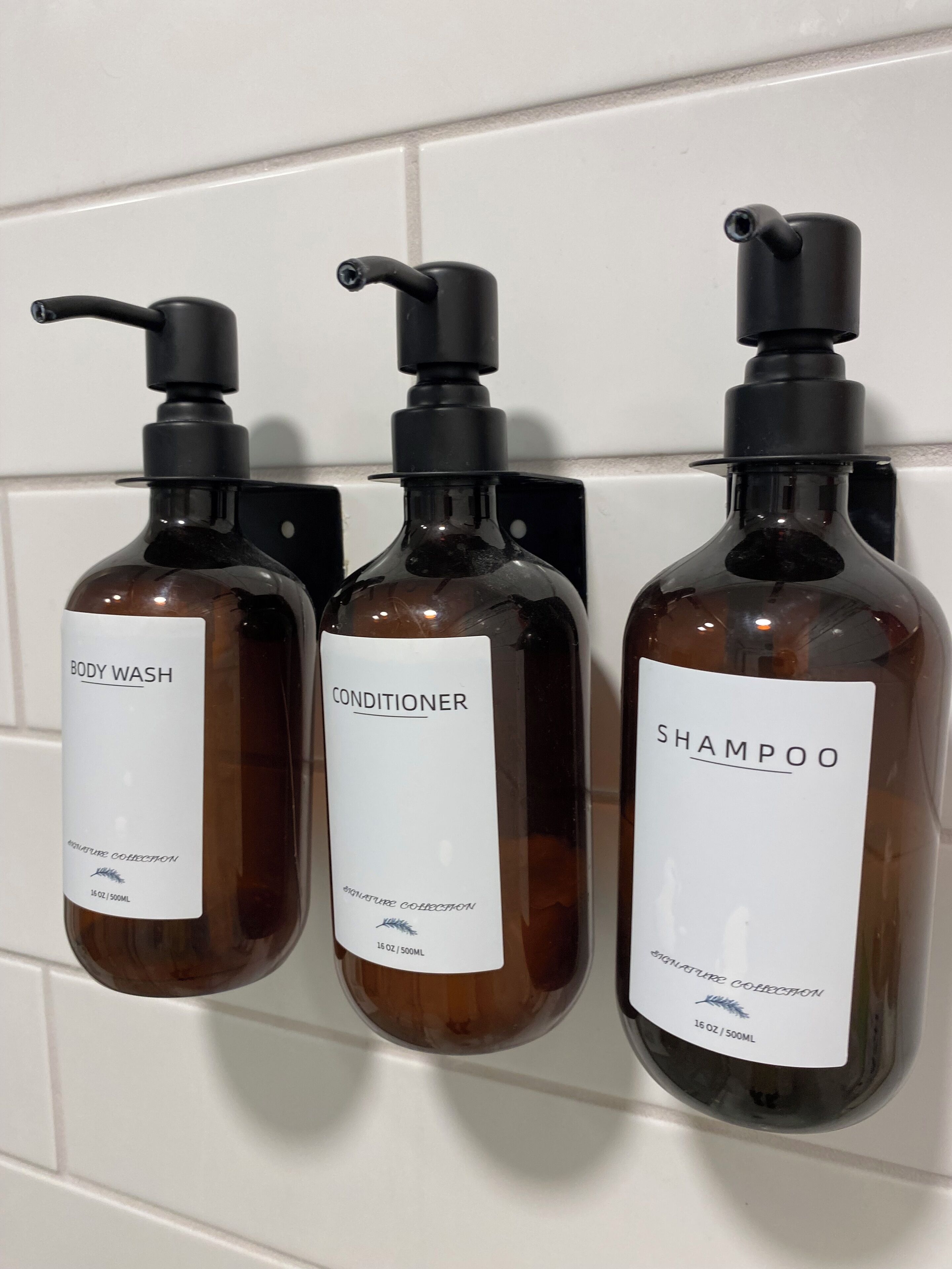 Tres botellas de gel de baño, acondicionador y champú sujetas a una pared de azulejos blancos.