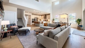 A spacious living area with a large grey sofa, a coffee table, and a dining table with chairs. A staircase leads to an upper level.