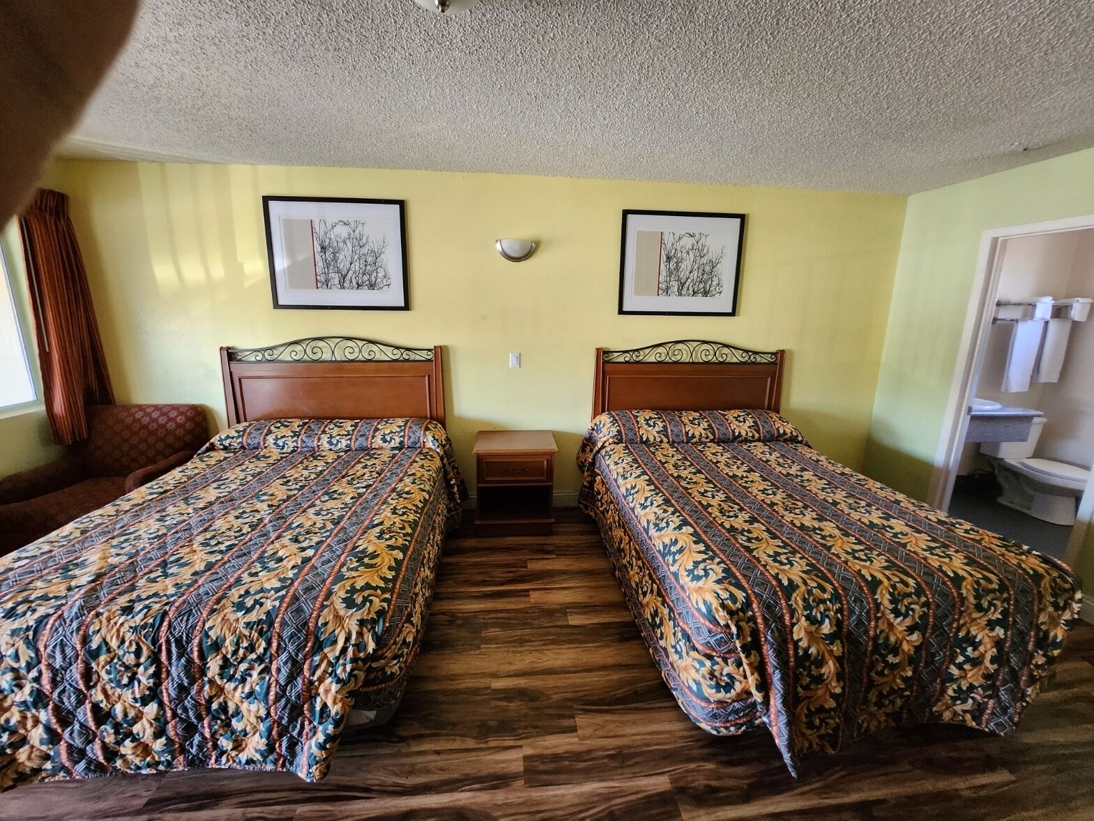 A room with two beds, wooden headboards, a small nightstand, and a window with curtains.