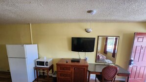 Een kleine kamer met een houten bureau, een witte koelkast, een magnetron, een televisie en een spiegel.