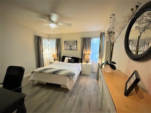 A bedroom with a bed, bedside tables, a desk, a chair, a ceiling fan, a mirror, and a potted plant. - Cozy Home near NOS Center/CSUSB (San Bernardino)
