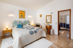 A bedroom with a bed, bedside tables, a nightstand, a mirror, and a bathroom visible through an open door.