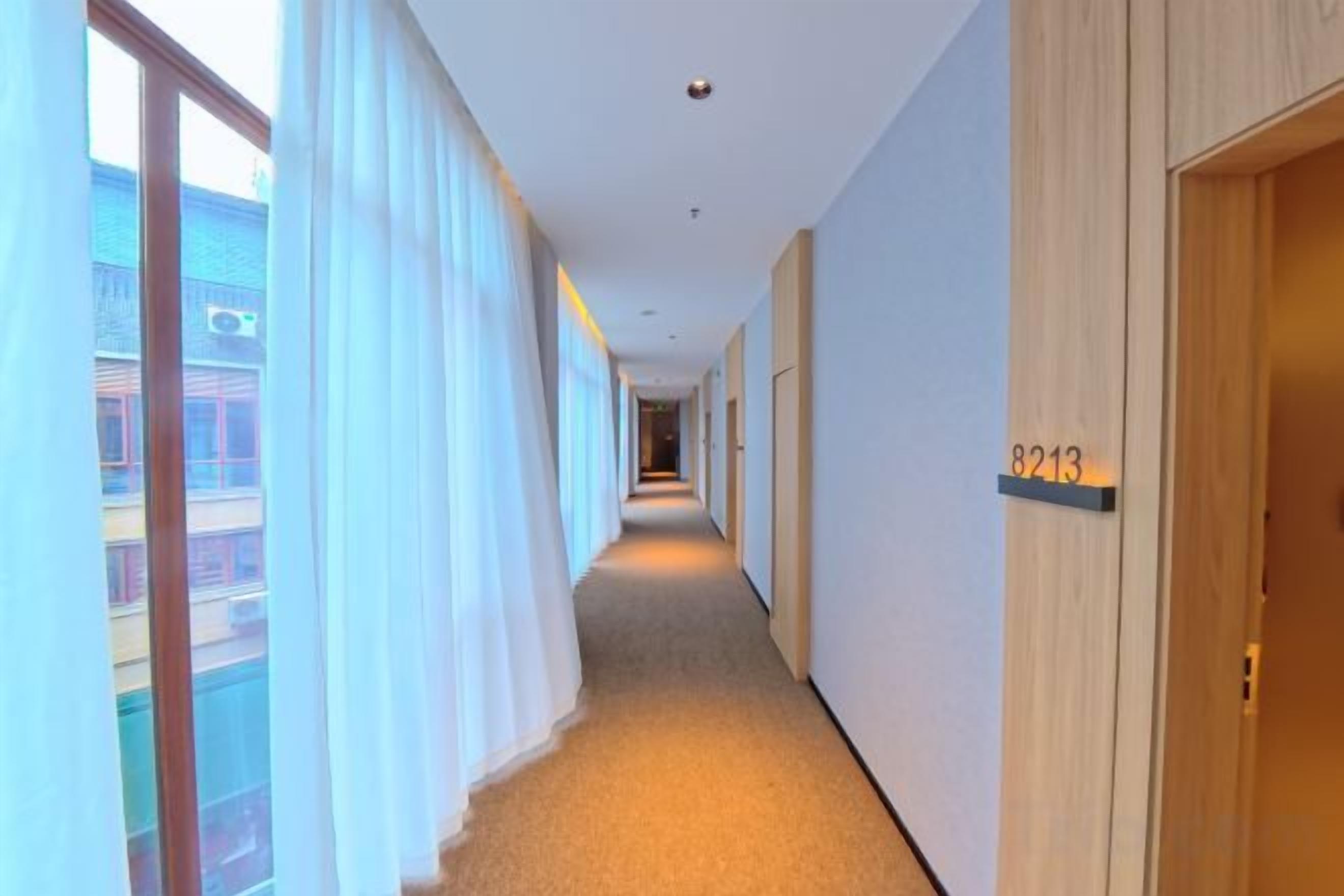 Un pasillo de hotel con piso alfombrado, una puerta de madera con número y una ventana con cortinas.