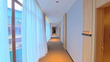 Un pasillo de hotel con piso alfombrado, una puerta de madera con número y una ventana con cortinas.