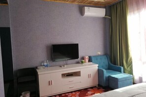 Une chambre d’hôtel dotée d’un téléviseur à écran plat fixé au mur, d’un mini-bar blanc, d’un fauteuil bleu et d’un lit recouvert d’une couvre-lit à motifs floraux.