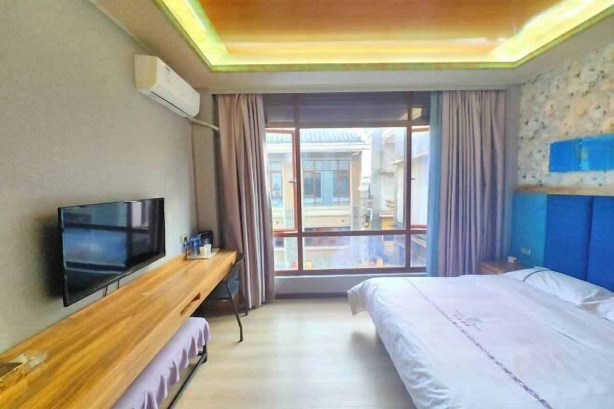 A hotel room with a bed, a TV on a wooden stand, a desk, and a window with curtains.