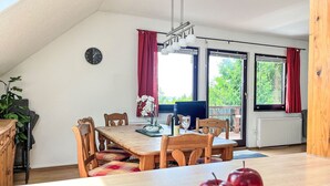 Zona pranzo con tavolo in legno, sedie, un orologio e vista sul verde circostante.