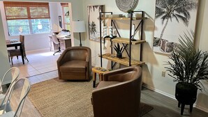 Una sala de estar con un sillón de cuero marrón, una mesa de centro de madera, una lámpara de pie, un estante en la pared con objetos decorativos y un cuadro grande de una palmera.