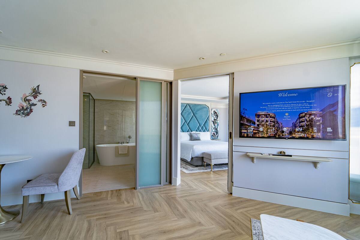 Een moderne hotelkamer met een groot bed, een televisie waarop een welkomstbericht staat, een badkamer met een bad en een zithoek met een stoel.