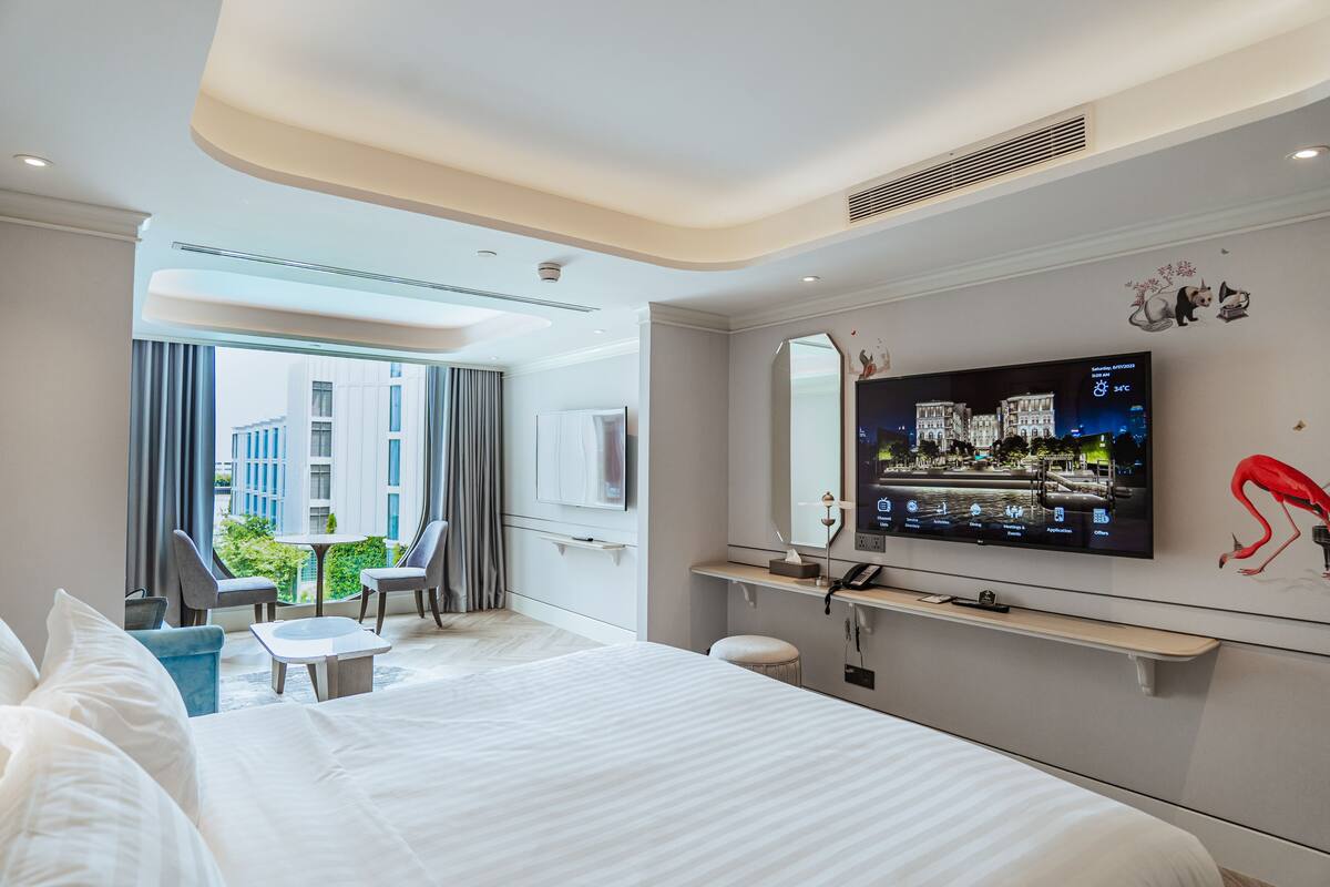 Een moderne hotelkamer met een groot bed, een televisie, een bureau en uitzicht op de stad door grote ramen.