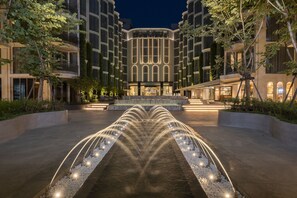 Un hôtel moderne doté d’un élément aquatique central, entouré de bâtiments bien éclairés et de verdure.