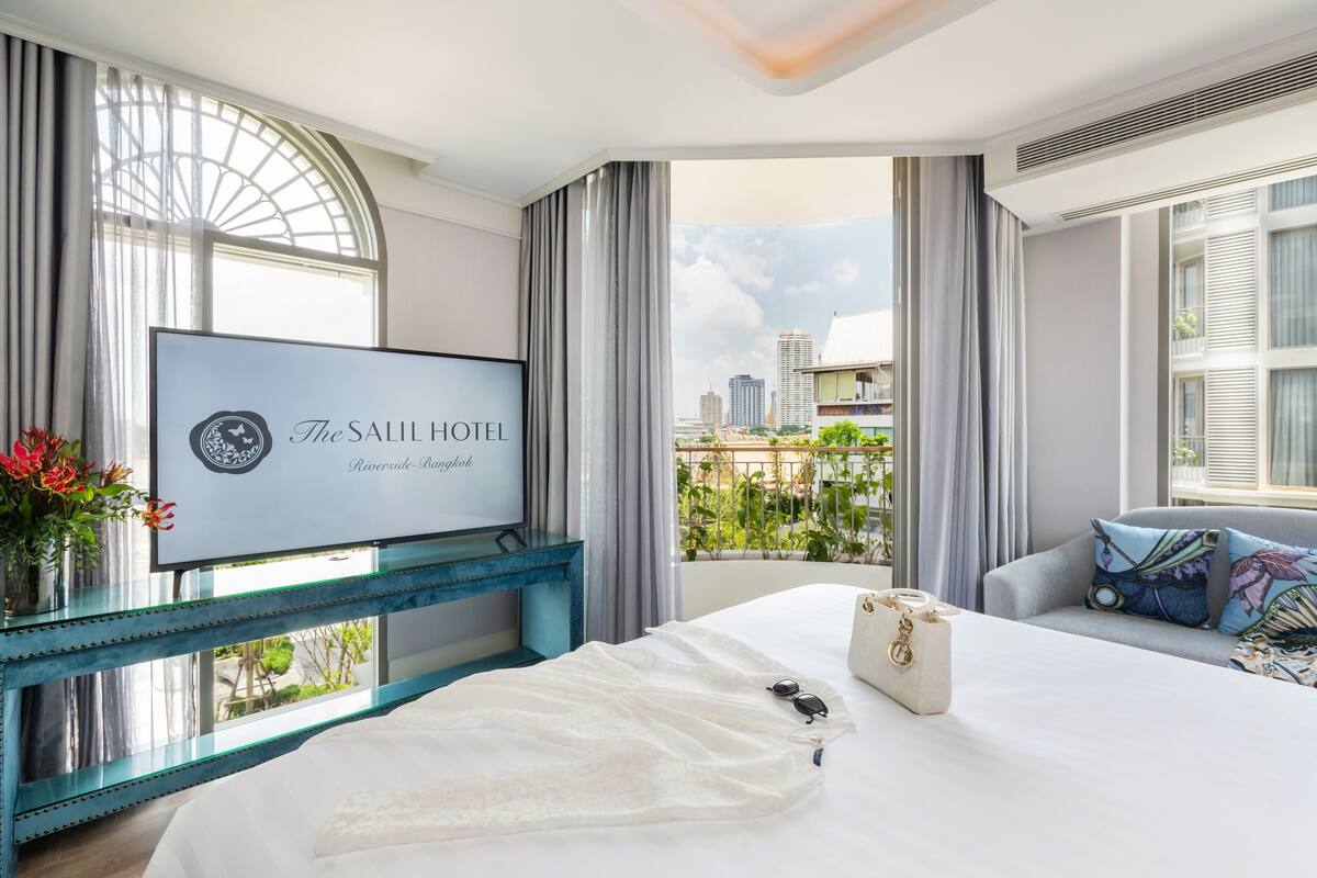 Een hotelkamer met een bed, een televisie bevestigd aan een blauwe console, uitzicht op de stad en een balkon met planten.