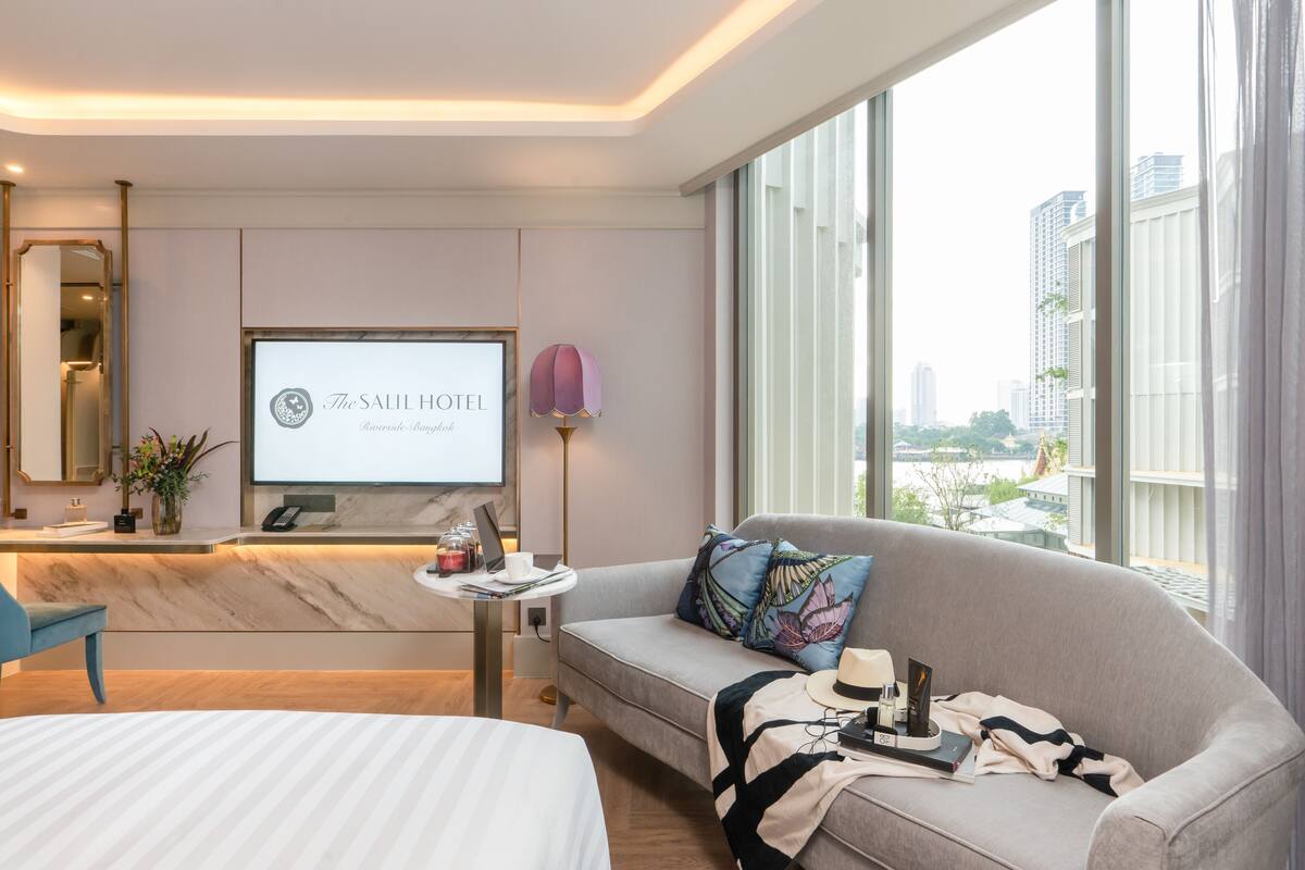 Een moderne hotelkamer met een bed, een sofa, een bureau met een laptop, een televisiescherm waarop de naam van het hotel staat, en een stadsgezicht door grote ramen.