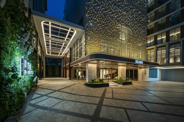 Una entrada de hotel moderna con fachada de vidrio, iluminación brillante y un área ajardinada con macetas.