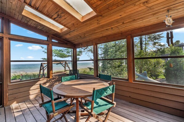 A wooden deck with a round table and chairs, large windows with a view of the sea, and a ceiling fan.