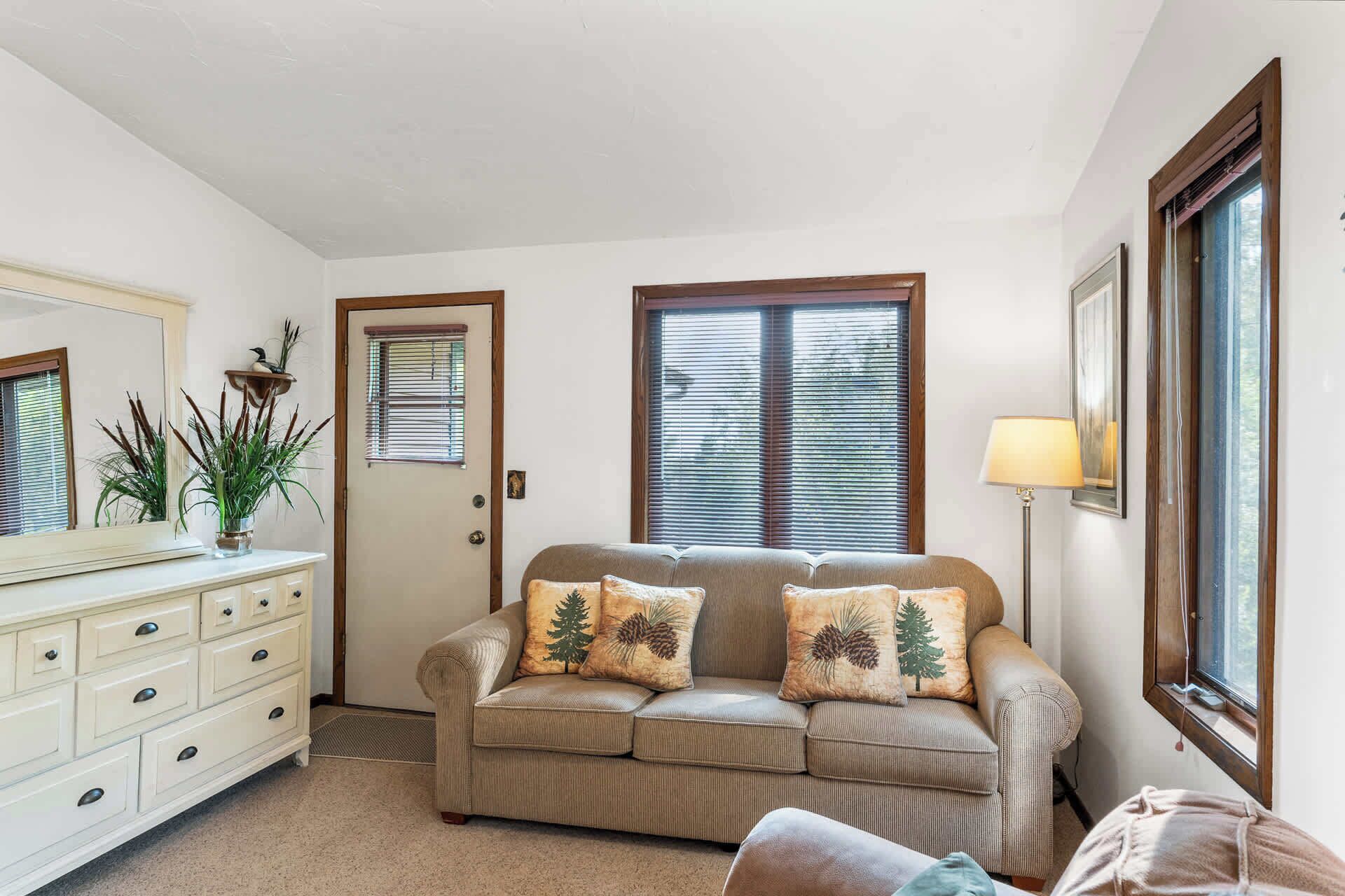 A living room with a beige sofa, two matching cushions, a white dresser, a mirror, and a lamp.