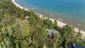 Vue aérienne d’une propriété située sur la côte, avec une plage, une forêt dense et une maison dotée d’un toit en pente.
