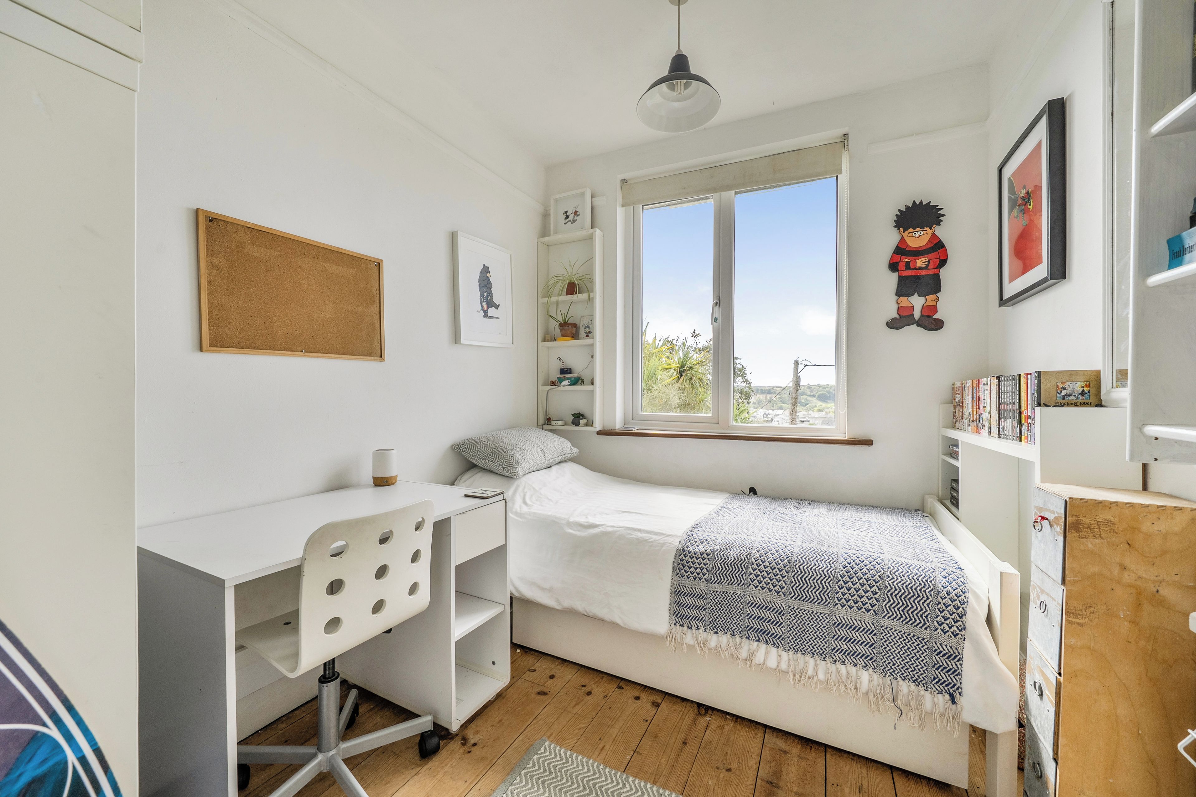 A bedroom with a single bed, a desk, a chair, a window, and a shelf with books.