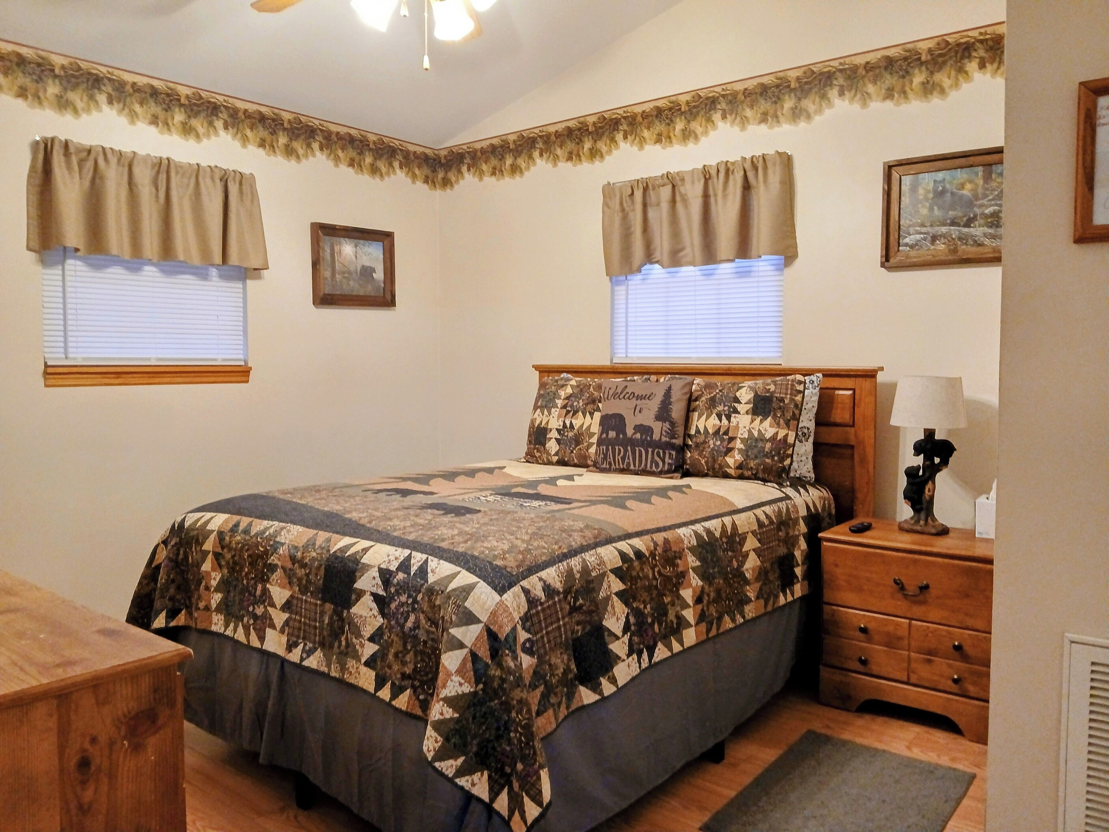 Une chambre à coucher comprenant un lit, une table de chevet, une commode et deux fenêtres avec des rideaux.