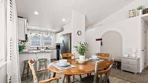 A dining area with a wooden table and chairs, a kitchen in the background, and a white wall with a clock. - Retro Caldwell Retreat | Backyard Hammocks | Sleeps 10 (Caldwell)
